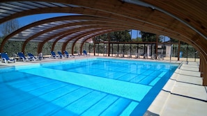 A large indoor swimming pool with clear blue water, surrounded by wooden beams and covered with a transparent roof. - Les Beaupins - Vacances ULVF (Saint-Denis-d'Oleron)