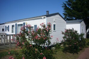 A white mobile home with blue shutters, surrounded by pink flowering plants. - Les Beaupins - Vacances ULVF (Saint-Denis-d'Oleron)