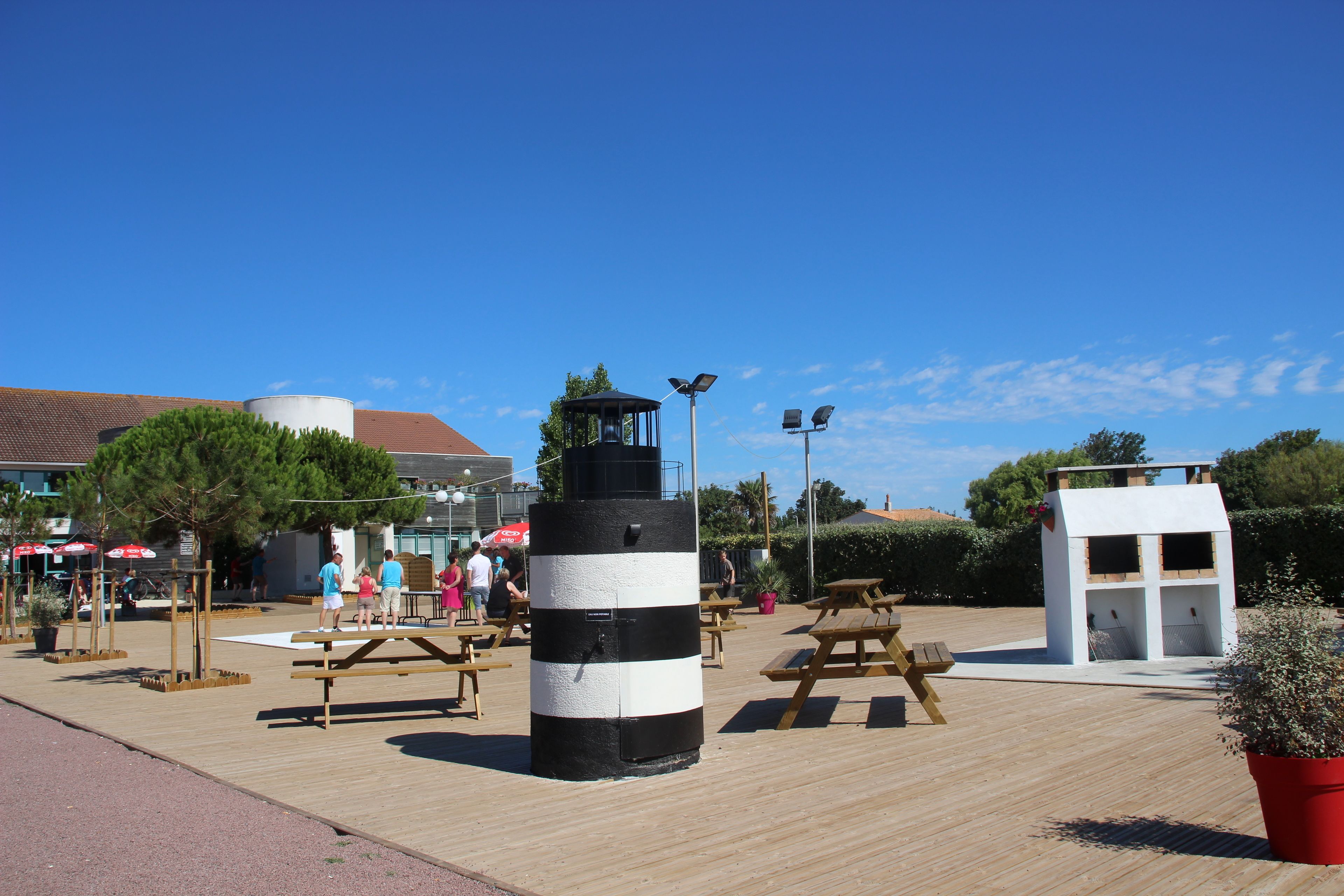 Una terraza de madera con mesas de picnic, un faro blanco y negro y un edificio blanco con una ventana pequeña.