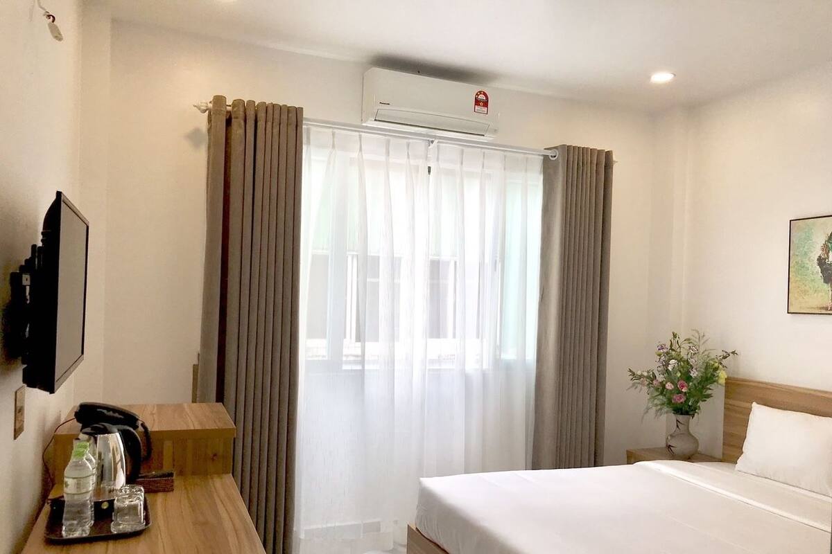 A hotel room with a bed, a desk with a phone and a tray of items, a vase with flowers, and a window with curtains.