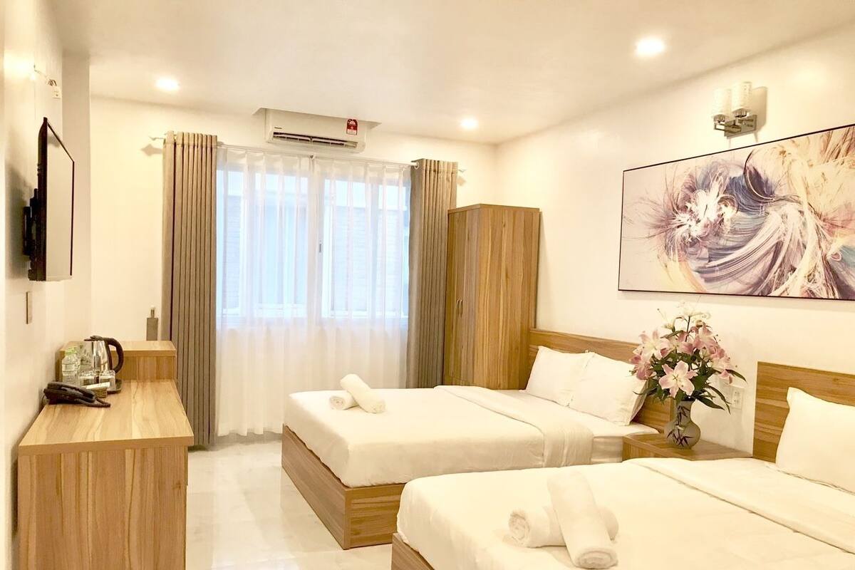 A hotel room with two beds, a wooden desk, a vase of flowers, and a painting on the wall.