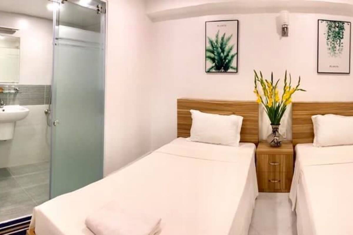 A hotel room with two beds, a glass shower enclosure, a bedside table with a vase of flowers, and framed artwork on the walls.