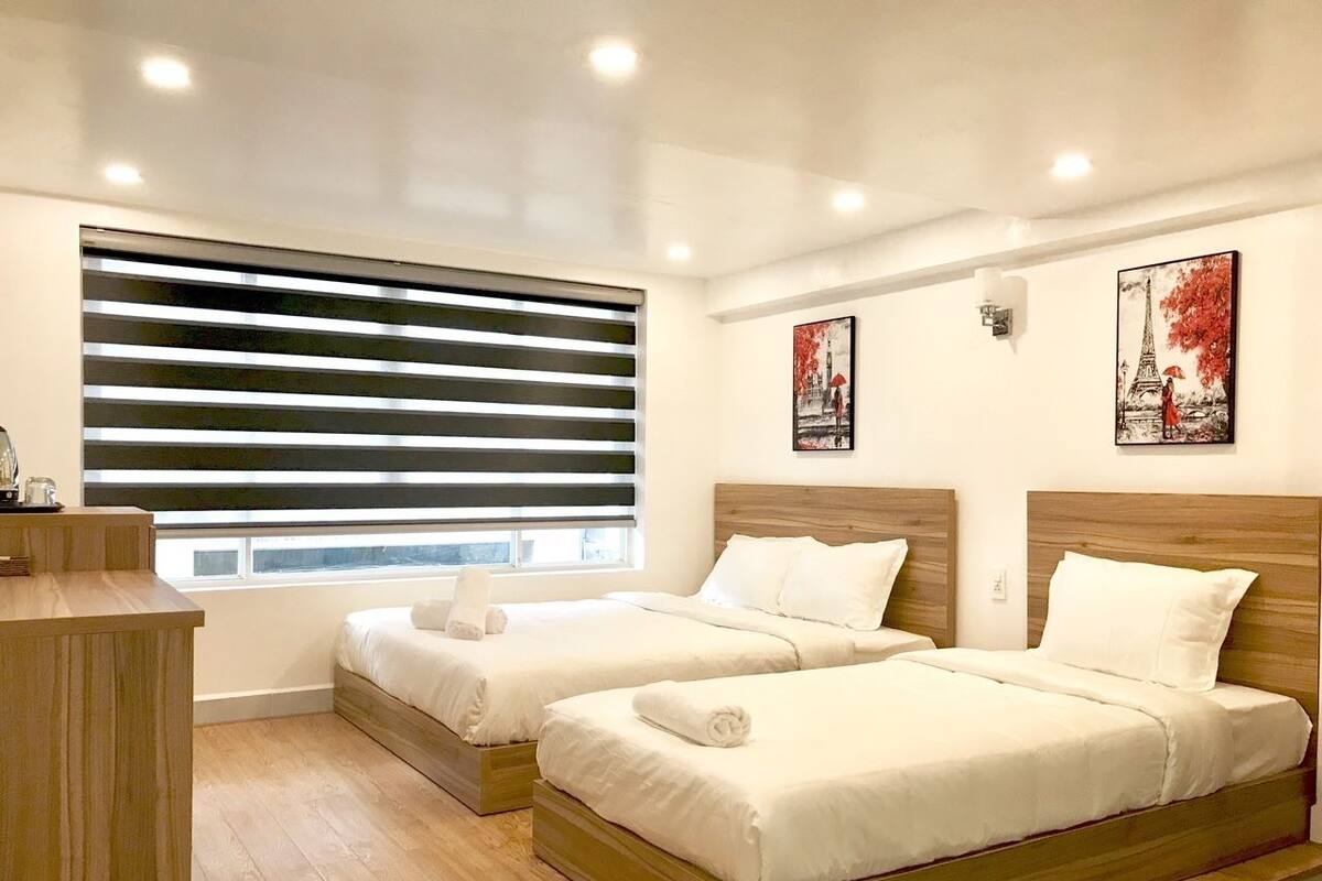 A hotel room with two beds, a wooden desk, and a large window with blinds.