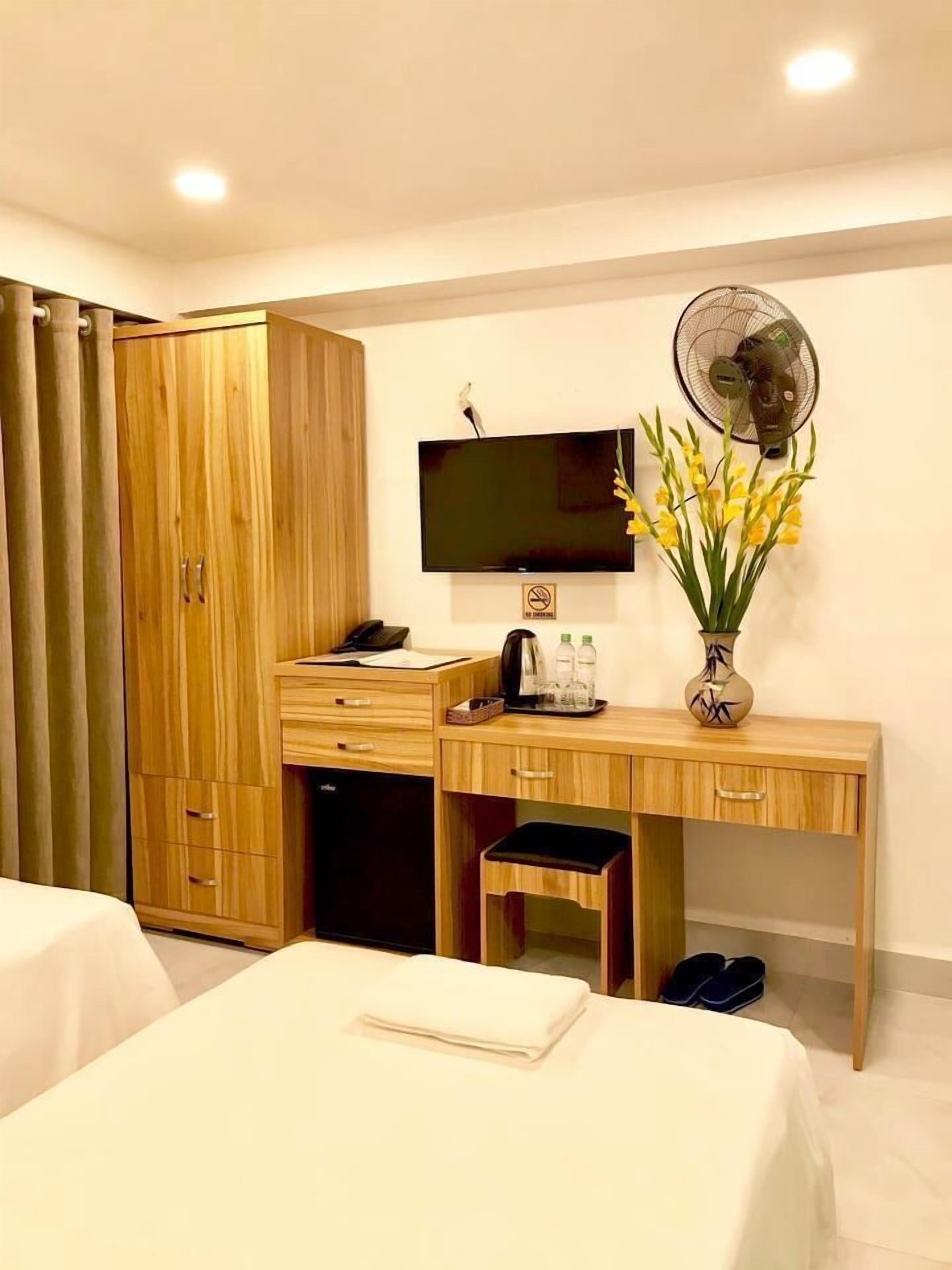 Quarto de hotel com guarda-roupa de madeira, uma escrivaninha com televisão de tela plana, um vaso com flores amarelas, um ventilador e um par de chinelos.