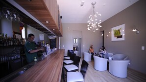 Zona de bar moderna con mostrador de madera, taburetes y un área de estar con sillas blancas.