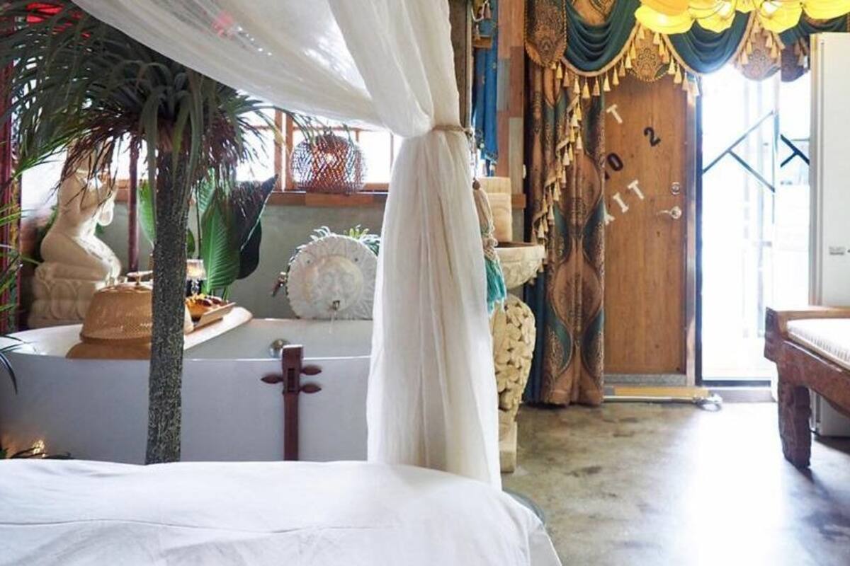 Um quarto com uma cama, uma mesinha, uma estátua e uma porta com número.