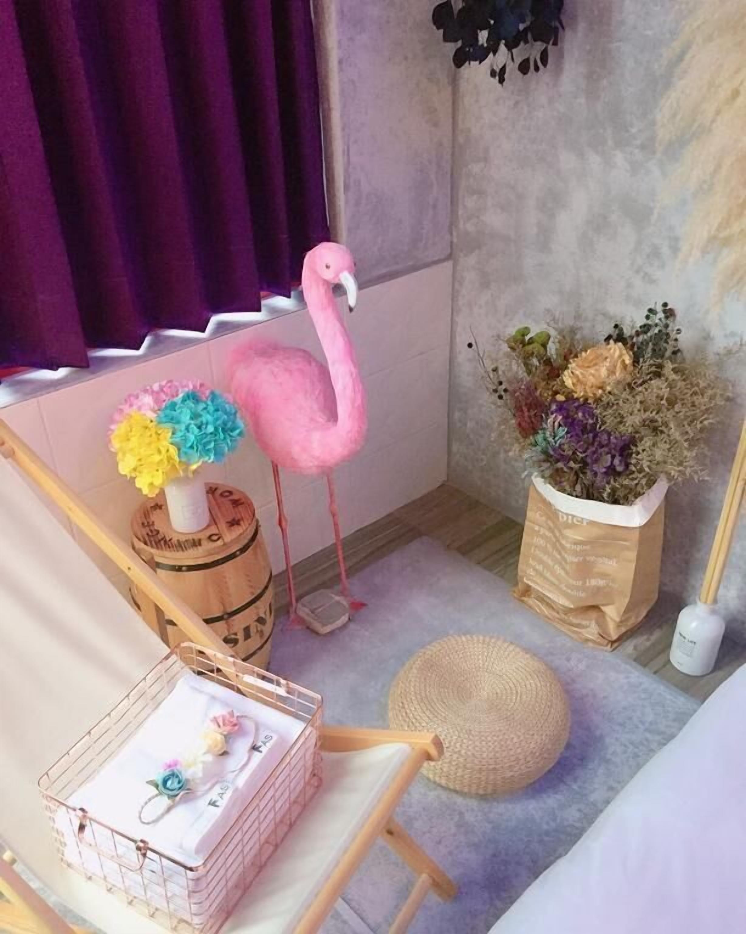 A room with a pink flamingo, a wicker basket, a wooden chair, a woven mat, and a potted plant with flowers.