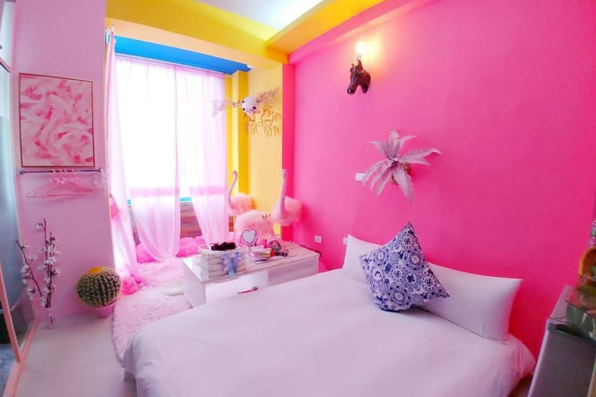 Um quarto vibrante com parede rosa, uma cama com almofadas brancas e azuis, uma mesa de cabeceira com livros e uma luminária, e uma janela com cortinas finas.