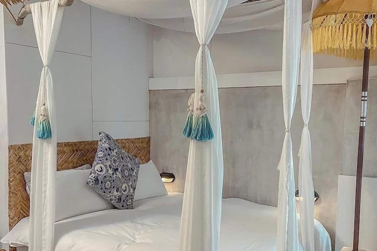 Uma cama de dossel com dossel, roupa de cama branca, borlas decorativas, uma almofada estampada e cabeceira de madeira.