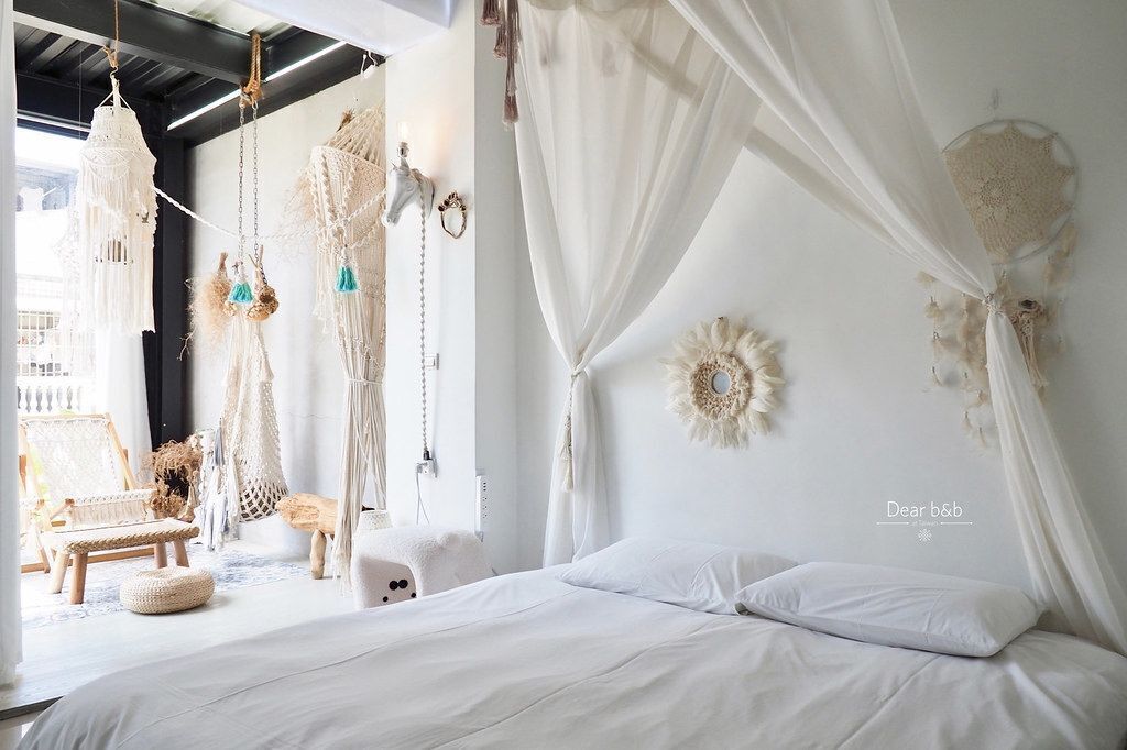 Un dormitorio con decoración blanca, una cama, adornos colgantes de macramé y un atrapasueños en la pared.