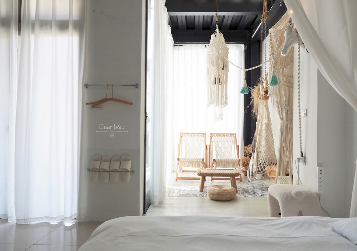 A modern bedroom with a bed, a hanging macrame chandelier, and a view of a room with two wicker chairs and a wooden bench.