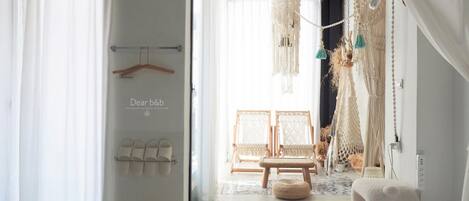 A modern bedroom with a bed, a hanging macrame chandelier, and a view of a room with two wicker chairs and a wooden bench.