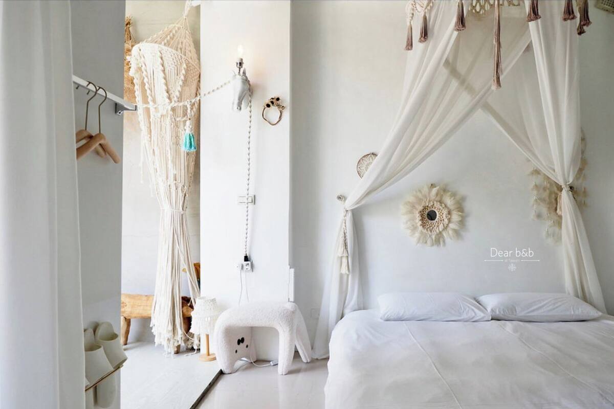 Quarto com cama, um pendente de macramé, um banquinho pequeno e uma luminária na parede.