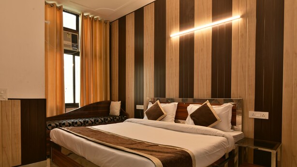 A modern hotel room with a large bed, a bedside table, a window with curtains, and a wall-mounted air conditioning unit. - Greno House (Greater Noida)