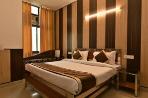 A modern hotel room with a large bed, a bedside table, a window with curtains, and a wall-mounted air conditioning unit. - Greno House (Greater Noida)