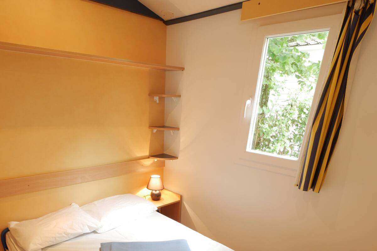 A single bed with a white mattress and a folded gray blanket, a bedside table with a lamp, and a window with curtains.