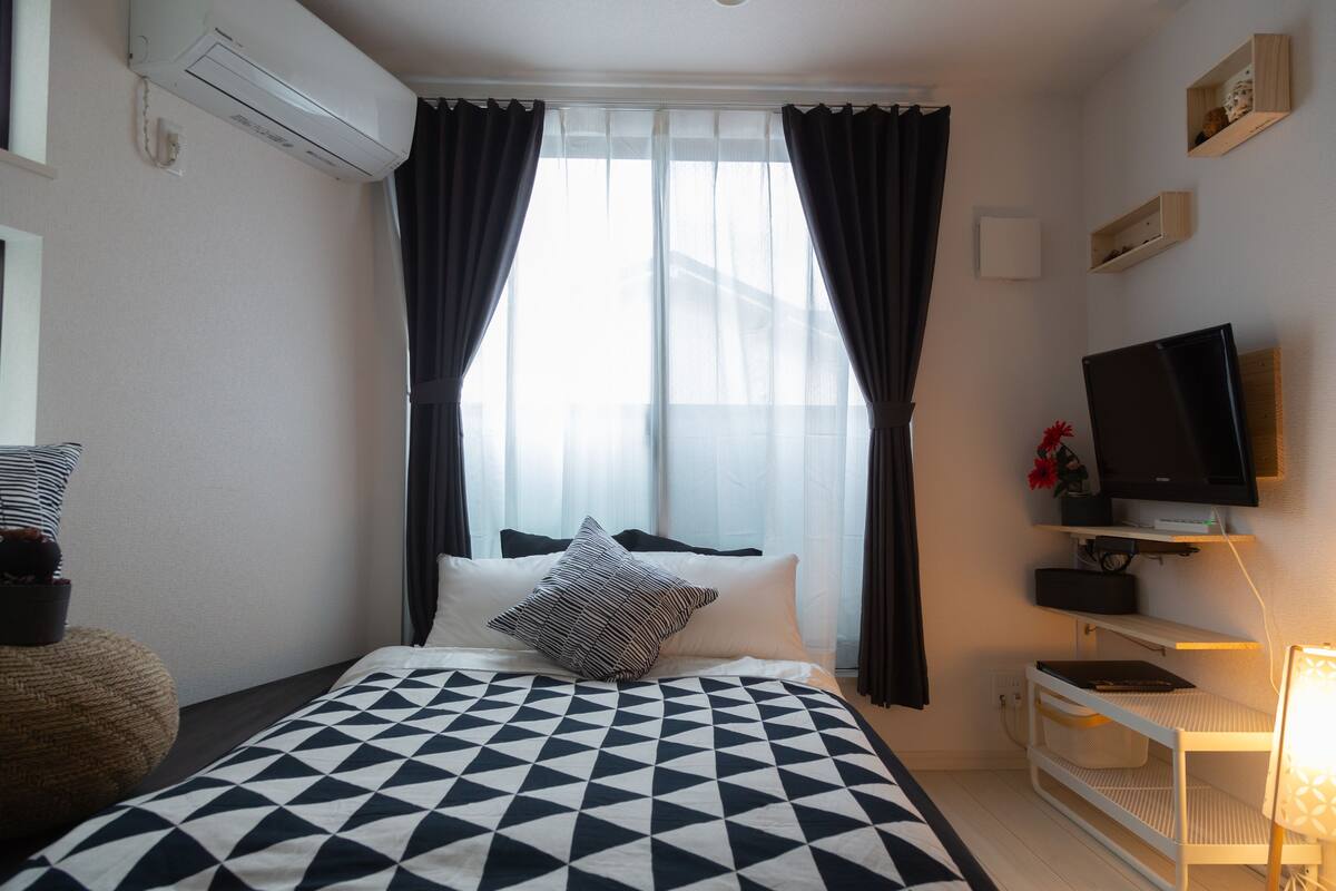 Hotellihuone, jossa on sänky, verhot, televisio ja pieni pöytä, jossa on lamppu.