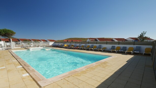 Une piscine rectangulaire à l’eau d’un bleu limpide, entourée d’une terrasse carrelée et bordée d’une rangée de chaises longues jaunes et bleues.