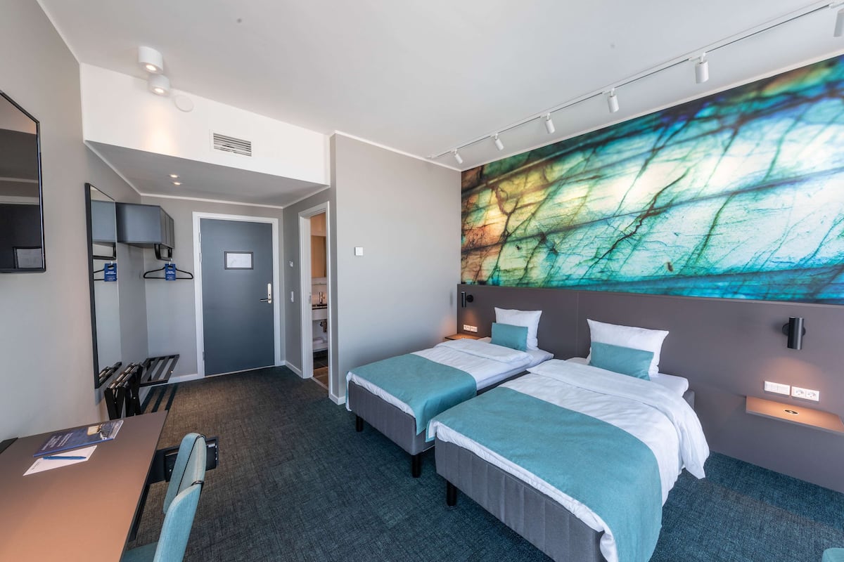 Ein Hotelzimmer mit zwei Betten, einem Schreibtisch, einem Stuhl, einem großen abstrakten Gemälde an der Wand und einer Tür zu einem Badezimmer.