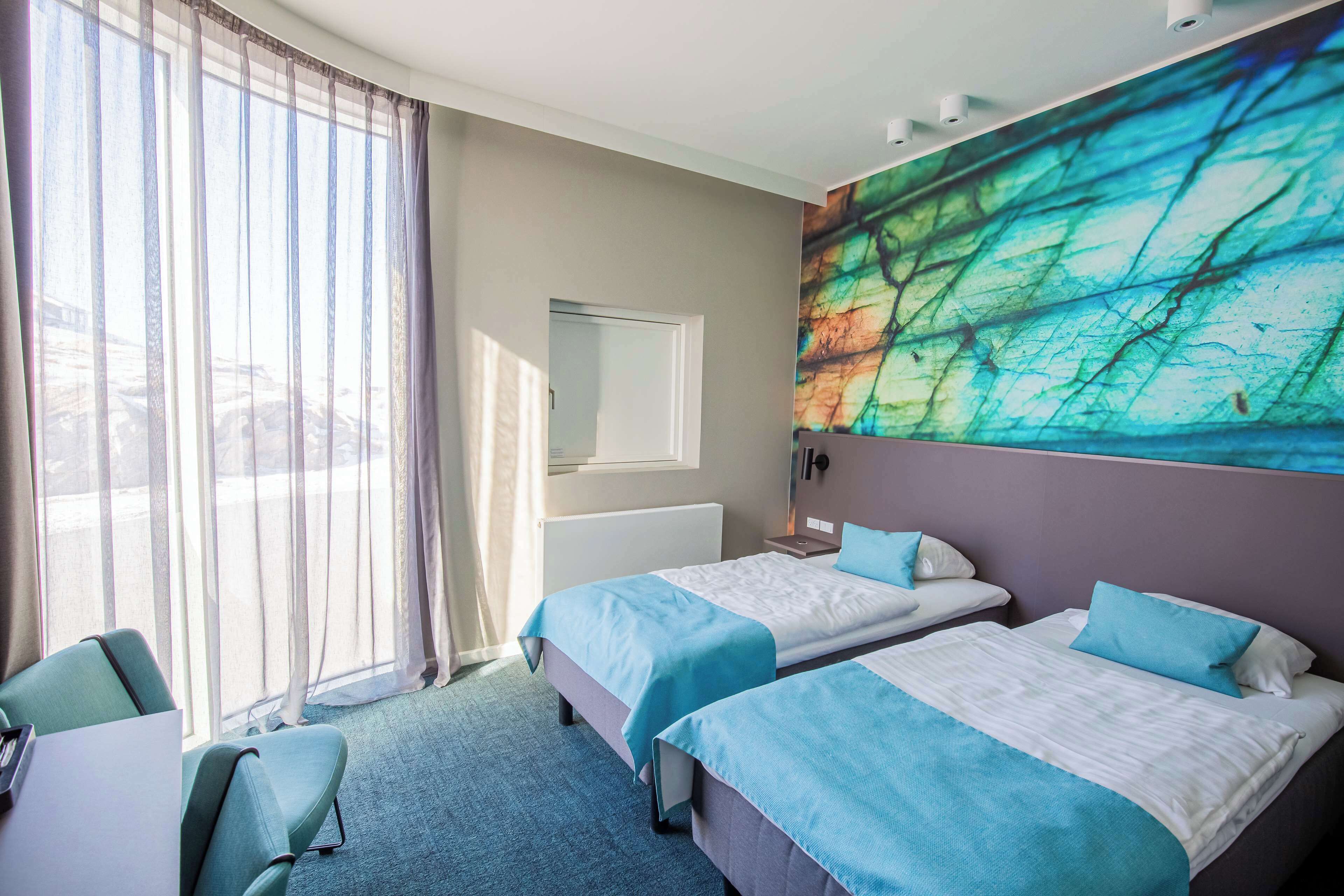 A hotel room with two beds, a large window with sheer curtains, a wall-mounted artwork, and a desk with a chair.