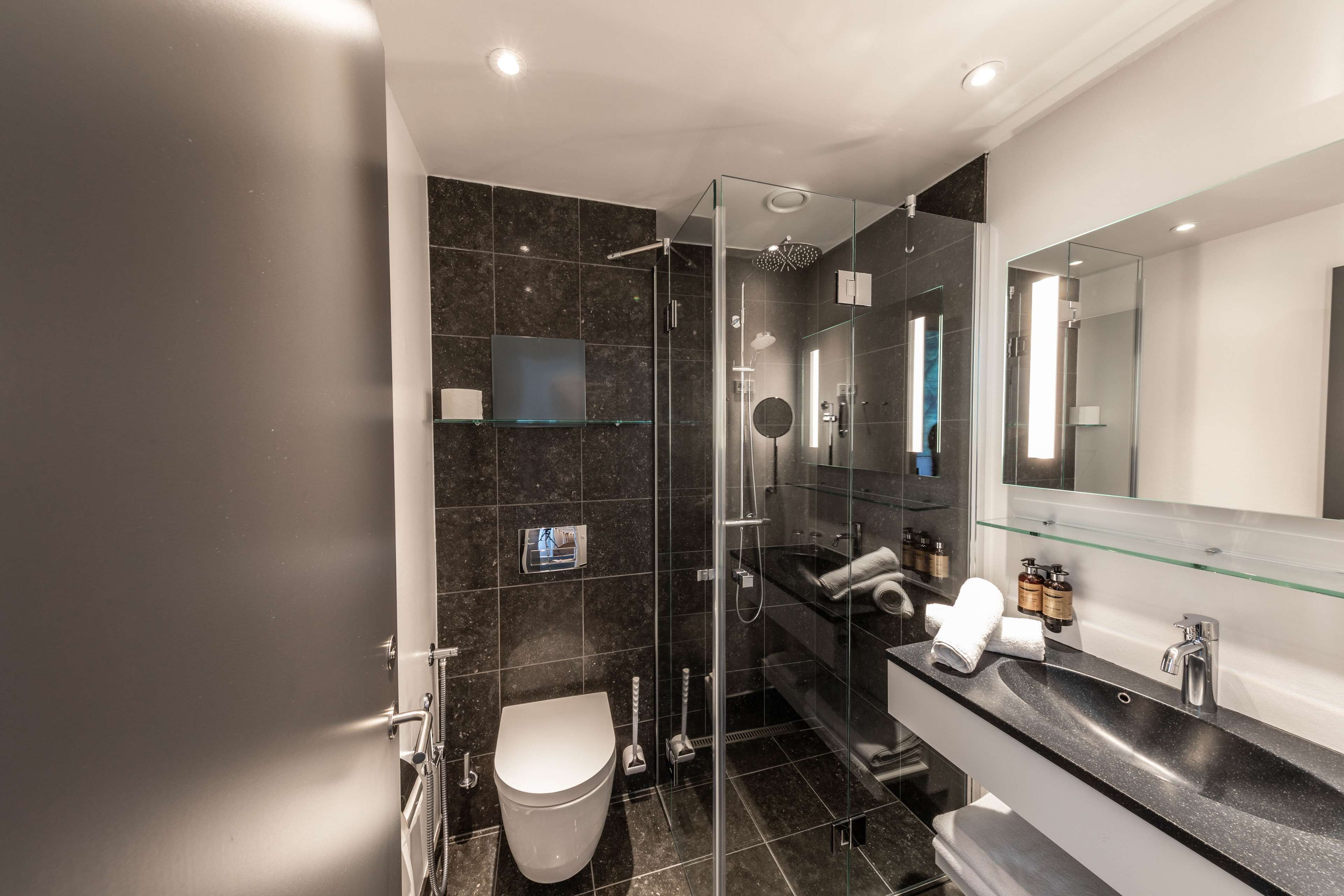 A modern bathroom with a glass-enclosed shower, a white toilet, a black sink with a glass countertop, and a large mirror above the sink.