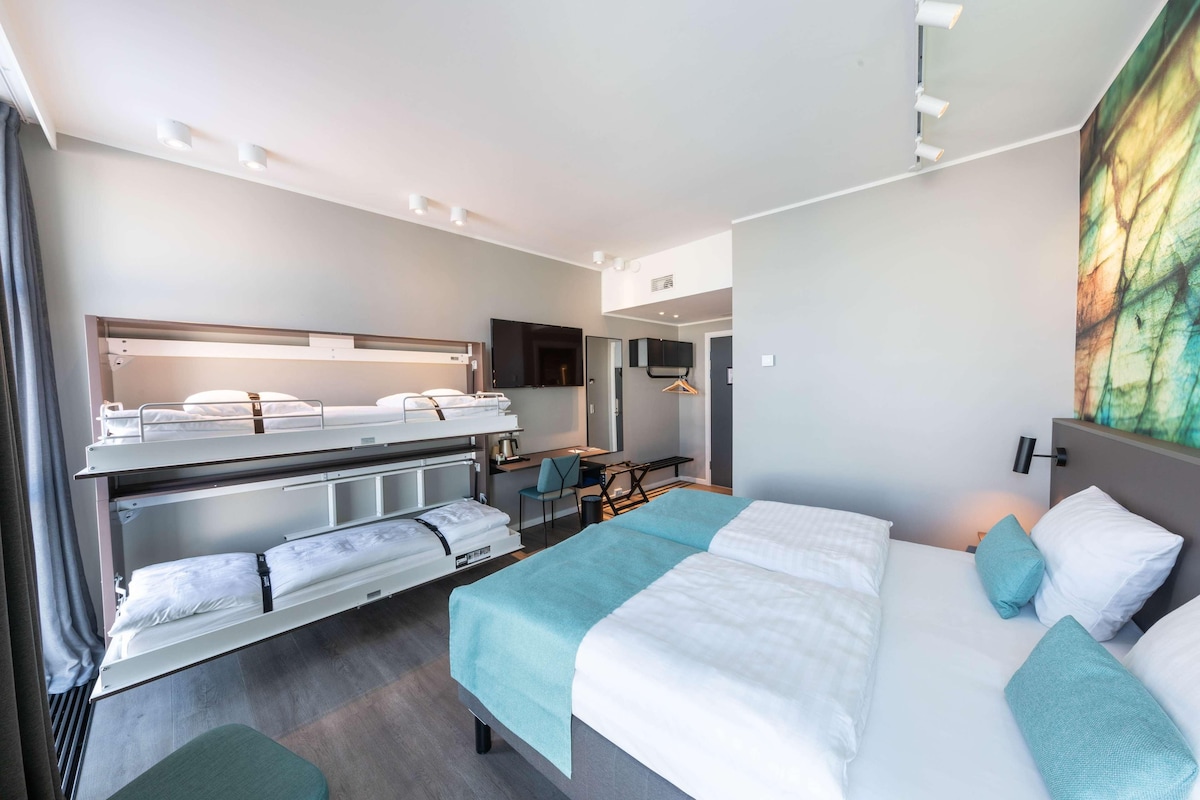 A hotel room with a bunk bed, a desk with a chair, a flat-screen TV, and a large window.