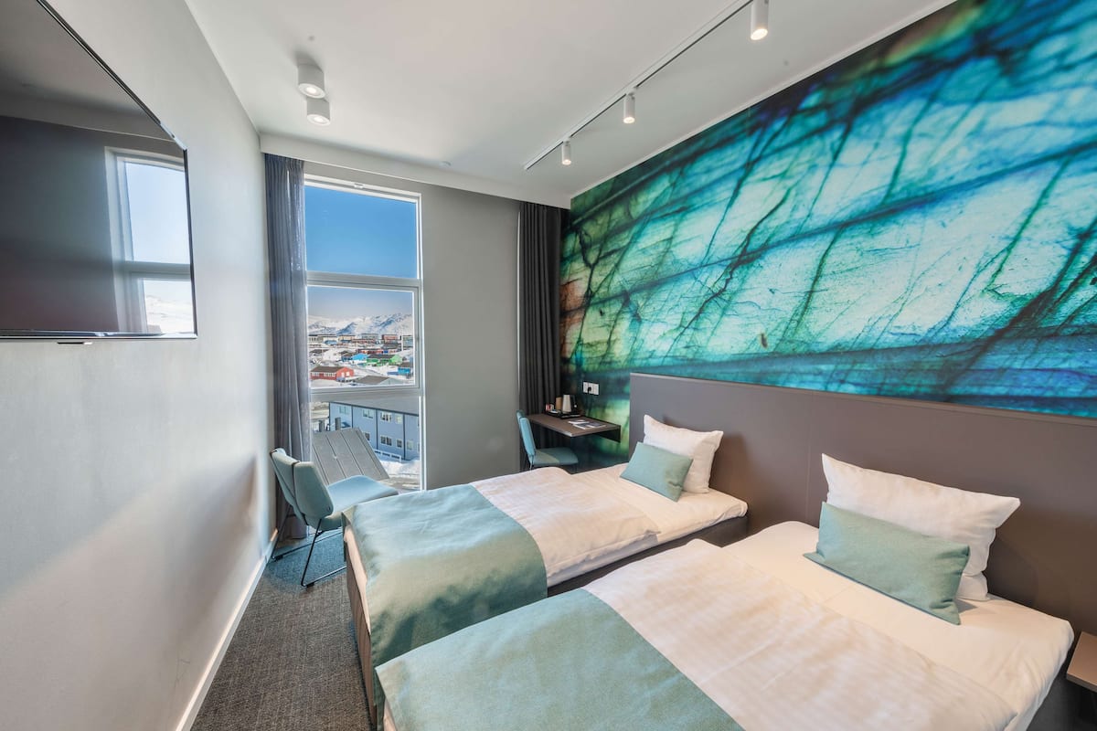 Ein Hotelzimmer mit zwei Betten, einem großen Fenster mit Blick auf eine Stadtansicht, einem Schreibtisch mit Stuhl und einem großen, abstrakten Gemälde an der Wand.