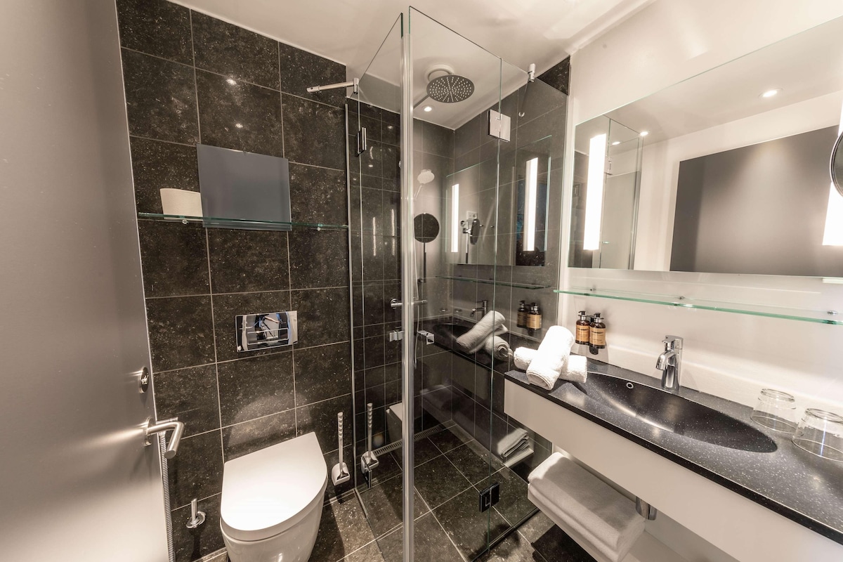 A modern bathroom with a glass-enclosed shower, a white toilet, a large mirror, and a black countertop with glass shelves.