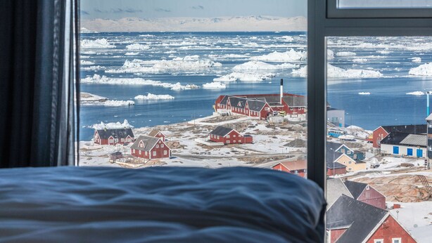 Une pièce dotée d’une grande fenêtre donnant sur un village côtier, avec ses maisons rouges et les icebergs qui flottent dans l’eau.