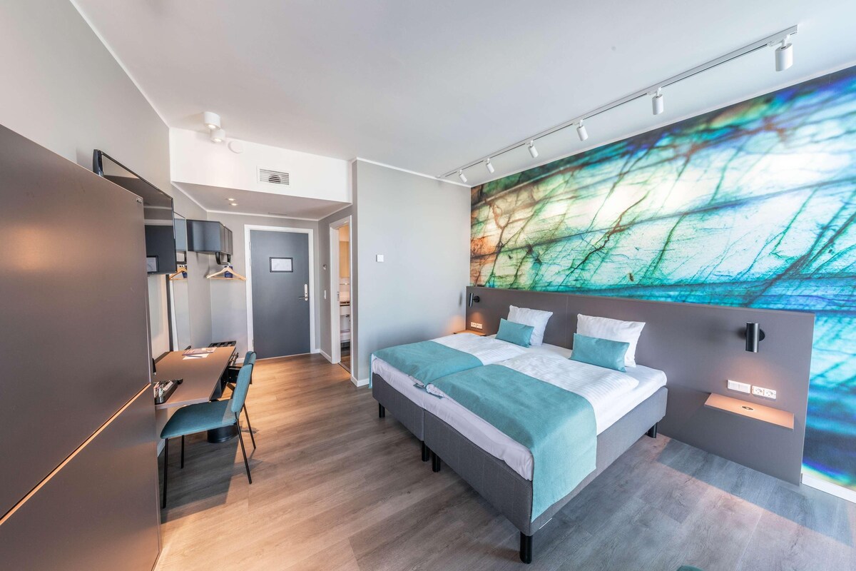 Une chambre d’hôtel moderne dotée d’un grand lit, d’un bureau et d’une fresque murale abstraite aux couleurs vives.