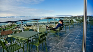 Un balcon avec des chaises et des tables en métal vert, donnant sur une ville côtière avec ses bâtiments et son pont.