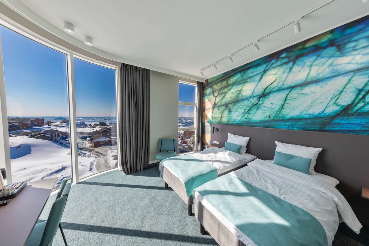 Ein Hotelzimmer mit zwei Betten, einem großen Fenster mit Blick auf die Stadt und einem modernen Kunstwerk an der Wand.