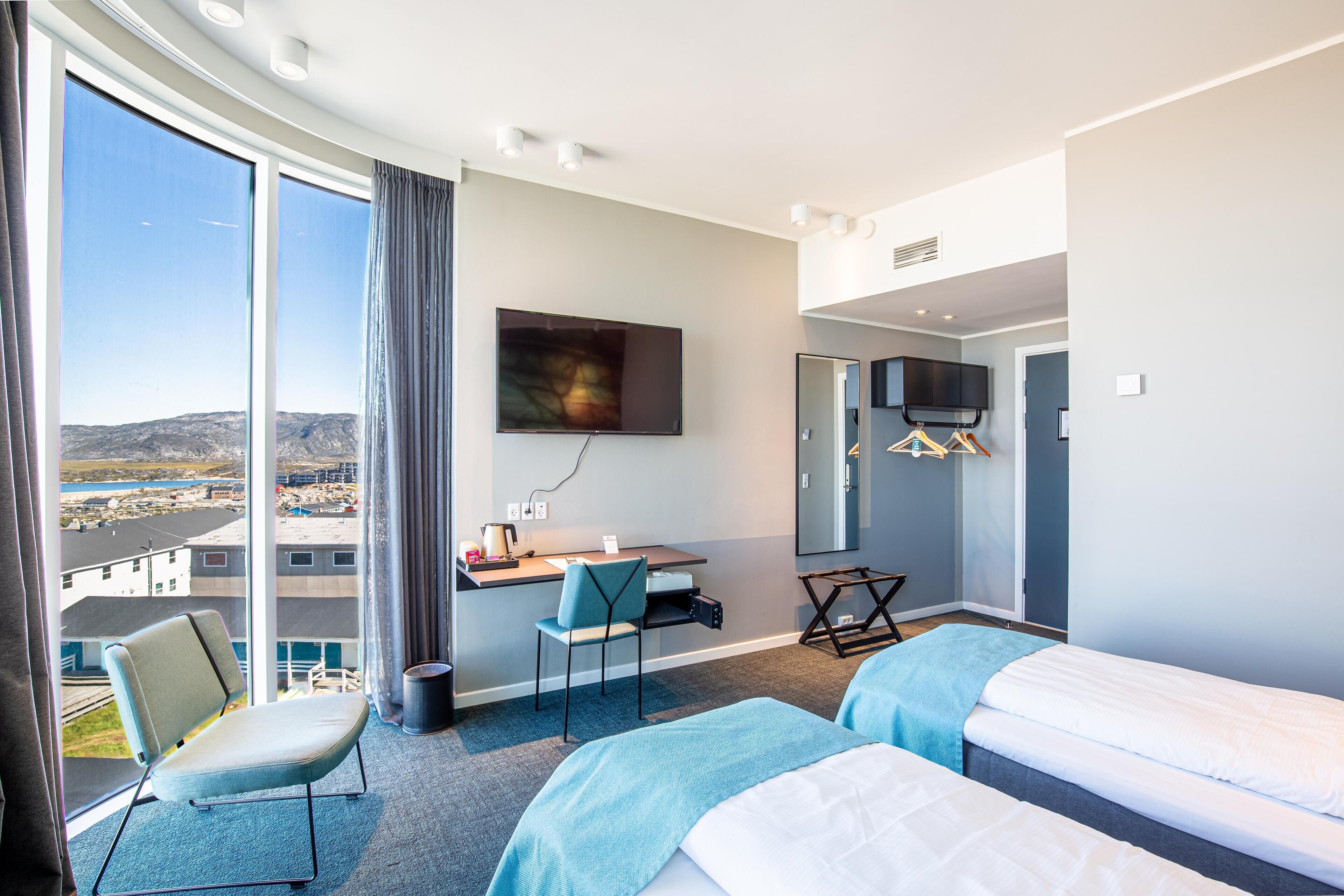 A hotel room with a large window, a desk, a chair, a bed, and a television.