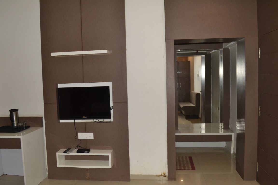 Une chambre moderne équipée d’un téléviseur fixé au mur, d’une petite étagère contenant des appareils électroniques et d’une porte-fenêtre donnant sur un autre espace.