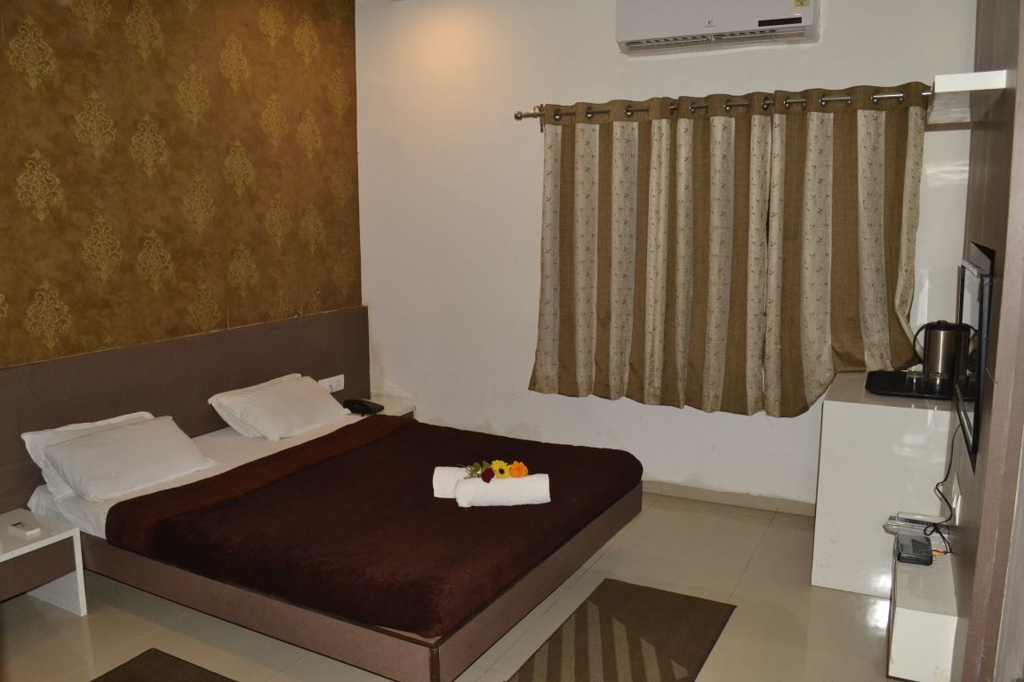 A hotel room with a bed, a nightstand, a desk with a kettle, and a curtain.