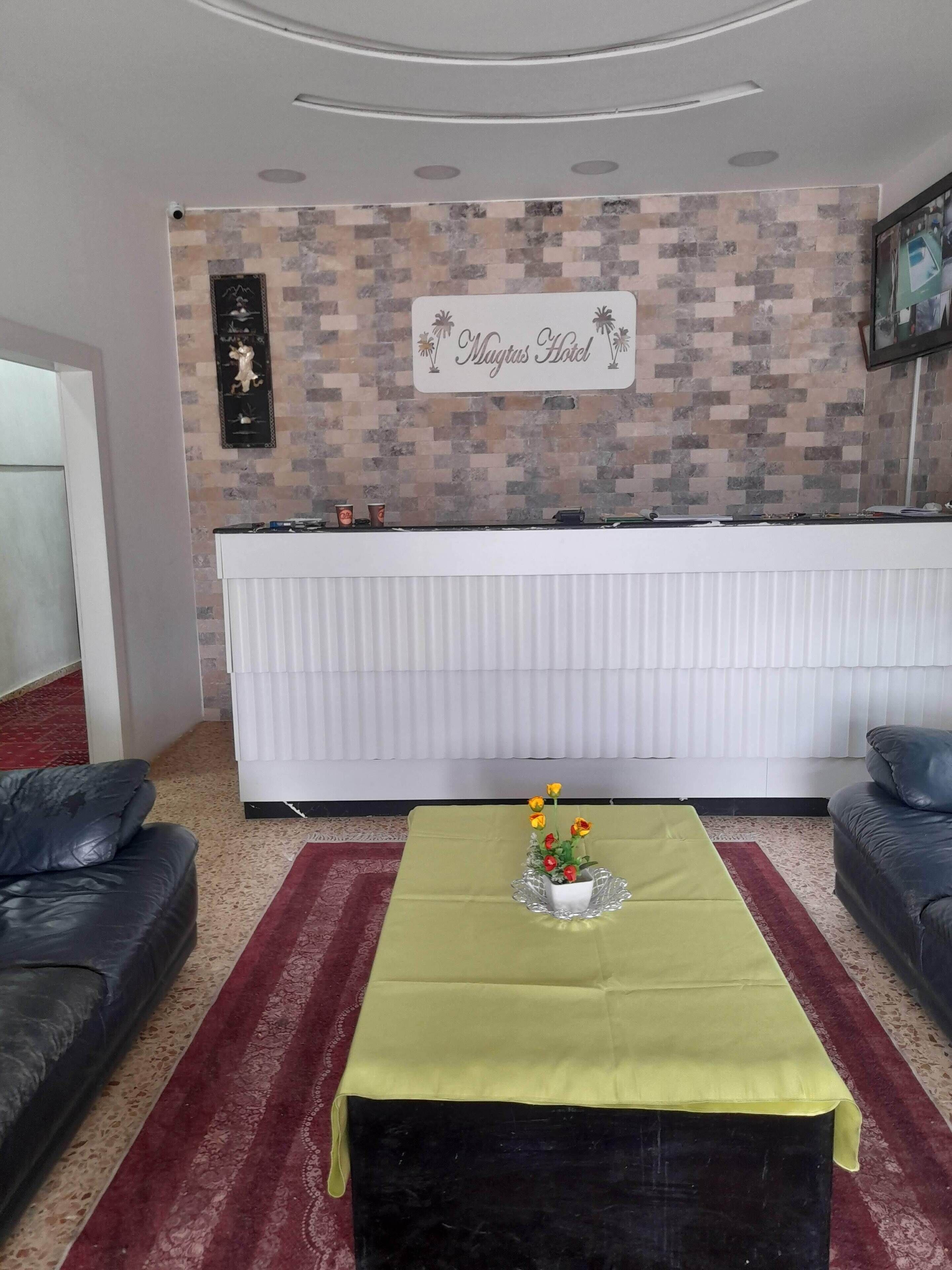 Un área de recepción con un letrero que dice "Mugtas Hotel", un mostrador de recepción blanco, una mesa verde con un florero y dos sofás negros.