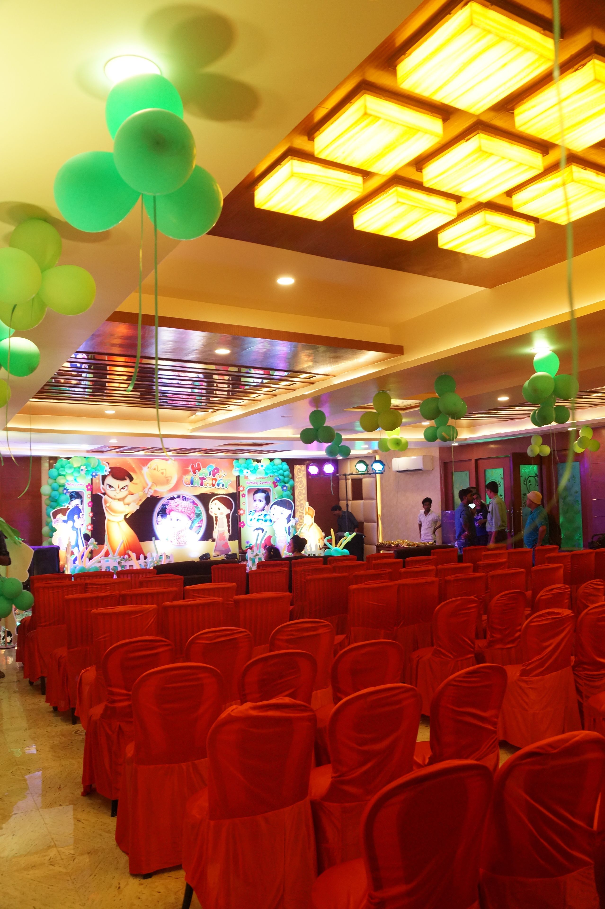 A banquet hall with rows of red-covered chairs, a stage with a backdrop, and green balloons.