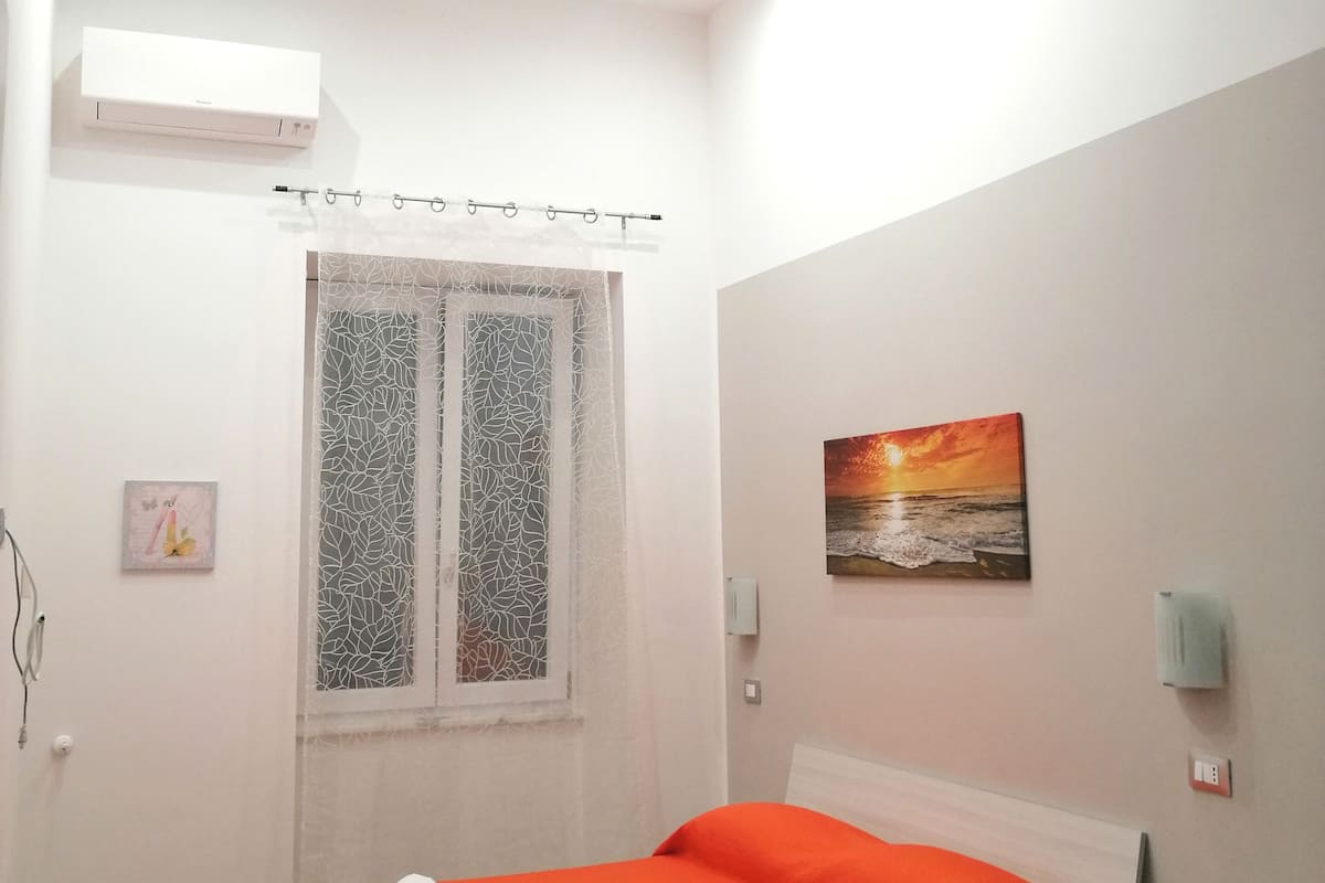 Ein Schlafzimmer mit Bett, einem Fenster mit dekorativer Vorhang und einem Wandbild, sowie einer Klimaanlage.