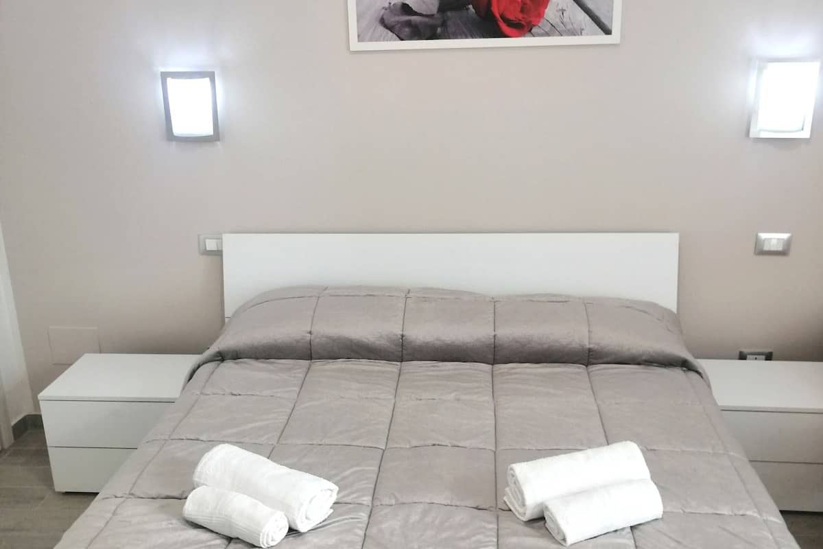 Ein ordentlich bezogenes Bett mit einer grauen Tagesdecke und zwei aufgerollten weißen Handtüchern, ein weißer Nachttisch, ein Beistelltisch und ein gerahmtes Bild an der Wand.