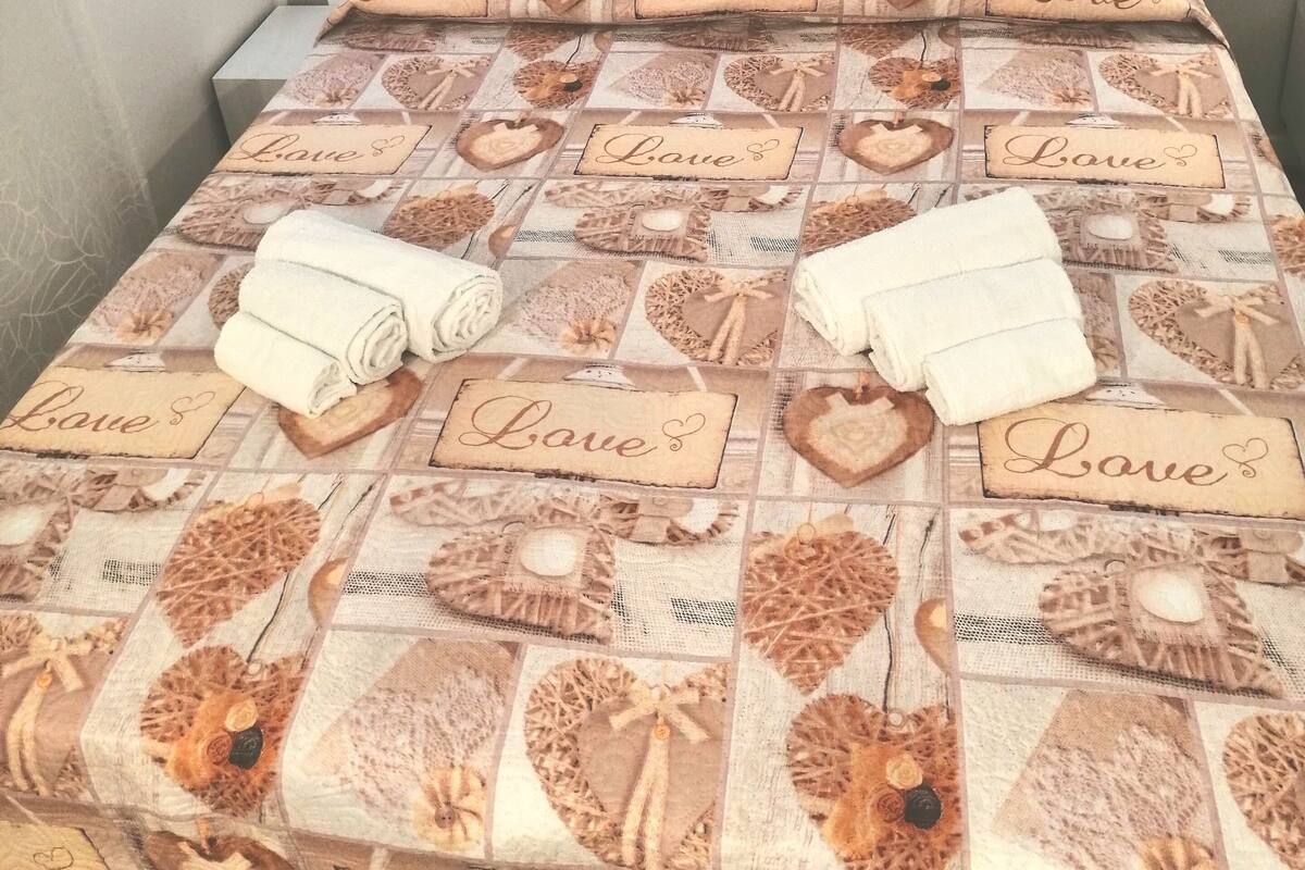 Ein ordentlich bezogenes Bett mit einem gemusterten Überwurf, der das Wort "Love" und Herzmotive zeigt, zwei an der Wand hängende Bilder und ein weißer Kopfteil.