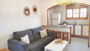 Een woonkamer met een grijze bank, een witte salontafel en een keuken met een koelkast en gootsteen.