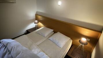 A neatly made bed with white linens and pillows, a wooden headboard, two bedside lamps, and a wall-mounted air conditioner.