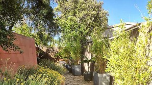 Un chemin de pierre bordé de plantes en pot et de fleurs jaunes, menant à un bâtiment avec un mur rouge et un toit recouvert de tuiles.