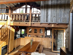 Un chalet rustique doté d’un coin repas en bois, d’une cuisine aux murs en pierre et d’une chambre à l’étage avec une rambarde en bois.