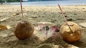 Zwei Kokosnussgetränke an einem sandigen Strand mit Blick auf ein ruhiges Meer und ferne Hügel.