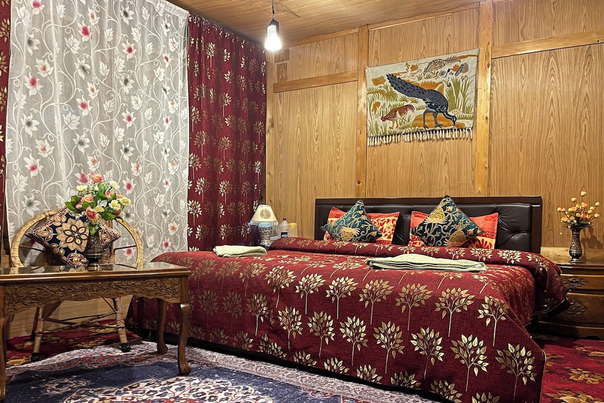 Un dormitorio con cabecera de madera, cortinas estampadas con motivos florales, cama con ropa de cama roja, un cojín decorativo, una mesita de noche con lámpara y un adorno en la pared.