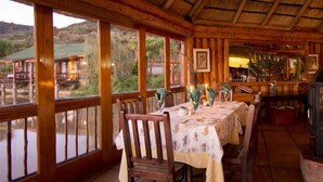 A wooden dining area with a table set for two, a fireplace, and large windows overlooking a natural landscape. - The Old Orchard (Greater Kokstad)