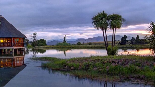 A lodge with a thatched roof, a dock extending into a calm lake, and a backdrop of mountains and palm trees under a twilight sky. - The Old Orchard (Greater Kokstad)