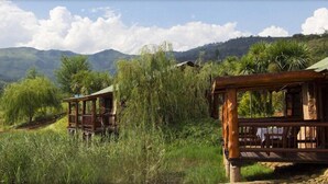 A rustic wooden cabin with a wooden deck, surrounded by lush greenery and mountains in the background. - The Old Orchard (Greater Kokstad)