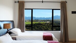 Ett hotellrum med två sängar, utsikt över berg och ett hus, och en lampa på vänster sida.